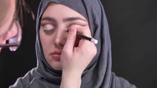 Portrait of female hands doing eye-makeup with white pencil for beautiful muslim woman in hijab on black background.