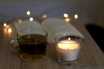 Scented candle, cup of tea, book and fairy lights. Cozy details at home. Selective focus.