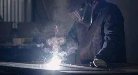 Professional welder in protective uniform with face mask, sparks fly apart, welding with an...