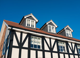 Three dormer windows in the roof of a Tudor style black and white English residential home against a blue sky. The house has sash windows and soft material string pull blinds