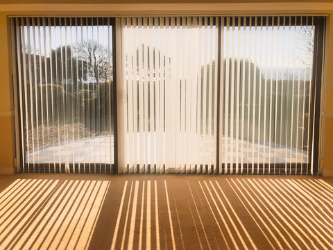 sunlight shining through full length white vertical blinds in front of three glass sliding french doors leading to a patio, garden and sea view, creating shadow stripes.