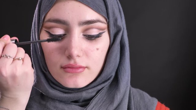Portrait of female hands doing make-up using mascara for attractive muslim woman in hijab on black background.
