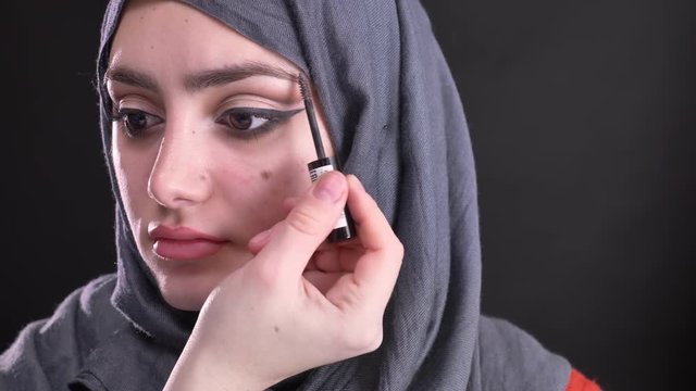 Portrait of female hands doing make-up and brushing eyebrows for attractive muslim woman in hijab on black background.