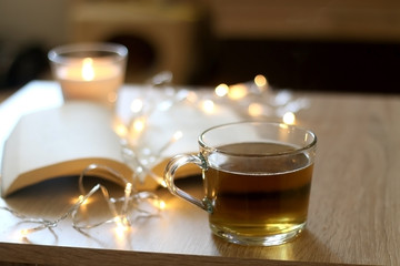 Scented candle, cup of tea, book and fairy lights. Cozy details at home. Selective focus.