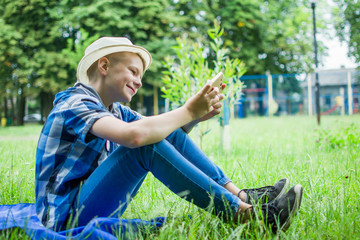 Lovely young guy wearing hat having a rest in a park with a phone. The guy makes a selfie and sits in social networks.