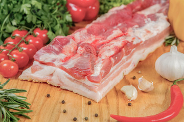 Fresh raw pork, meat is placed on a wooden board along with a branch of cherry tomatoes, lettuce, red peppers, chili, garlic and black pepper