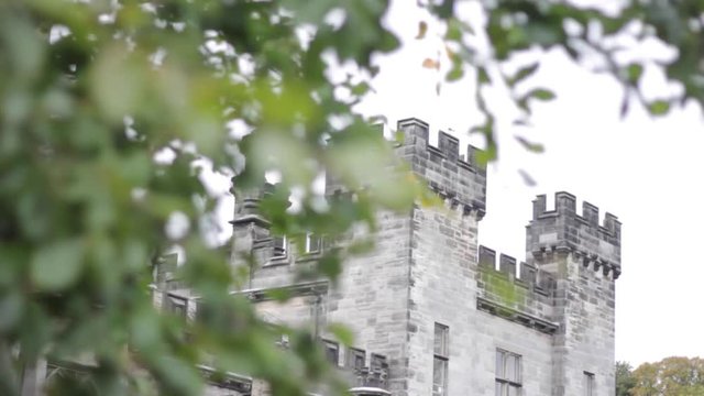 A close up on a castle turret pulls focus to the windy green tree leafs and branches in the foreground 