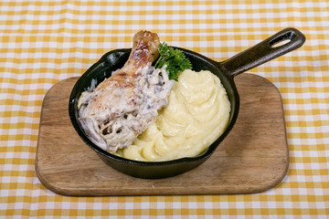 Fried chicken leg with mushroom sauce, paprika and garlic with mashed potatoes as a garnish and butter, served in a mini frying pan on a wooden board