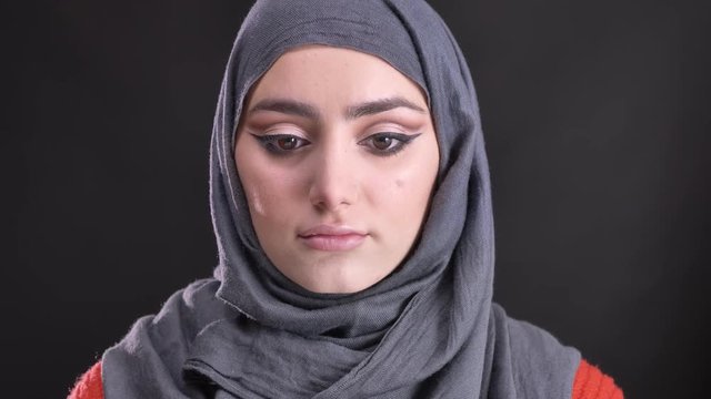 Portrait of beautiful muslim woman in hijab with bright and shiny make-up raises her eyes and watches calmly into camera on black background.
