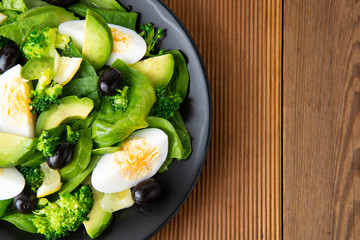 Avocado salad with broccoli, spinach, olives and boiled eggs in black plate, over wooden table. Healthy food. Avocado salad recipe.
