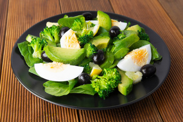 Avocado salad with broccoli, spinach, olives and boiled eggs in black plate, over wooden table. Healthy food. Avocado salad recipe.