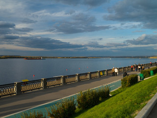 View of the city embankment with an openwork pig-iron protection, a green grass and the passing people in solar autumn evening against the background of the blue sky with clouds.