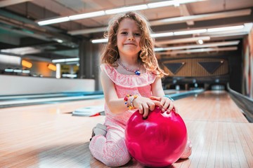 Portrait of a beautiful young girl with freckles smiling and holding a pink bowling ball, sitting on a bowling track.