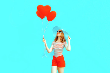 Happy smiling young woman with red heart shaped air balloons in summer straw hat and shorts on colorful blue background