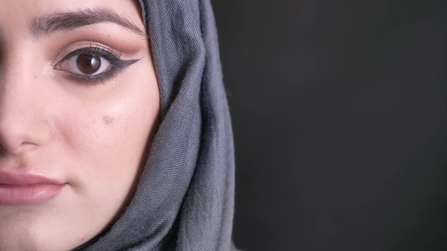 Close-up half-portrait of beautiful muslim woman in hijab with fashionable make-up watching calmly into camera on black background.