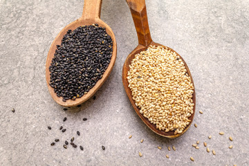 White and black sesame seeds in wooden spoons on stone background, top view, close up