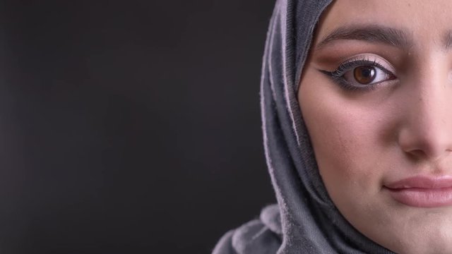 Close-up half-portrait of young muslim woman in hijab with fashionable make-up watching calmly into camera on black background.