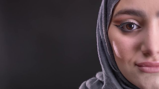 Close-up half-portrait of young muslim woman in hijab with fashionable make-up smiling into camera on black background