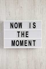 Modern board with text 'Now is the moment' on a white wooden background, overhead view. From above, flat lay, top view.