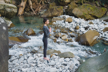 Young slim woman practicing yoga outdoors on stone shore of mountain river. Unity with nature concept. Girl standing in profile with prayer hands