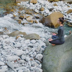 Young slim woman practicing yoga outdoors on stone shore of mountain river. Unity with nature concept. Meditation in easy pose.