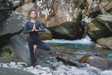 Young slim woman practicing yoga outdoors on stone shore of mountain river. Unity with nature...