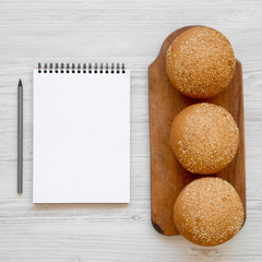 Fresh burger buns with sesame seeds on rustic wooden board, blank notepad on white wooden table, overhead view. Flat lay, from above, top view.