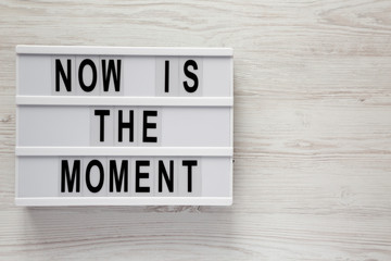 Modern board with text 'Now is the moment' on a white wooden table, top view. From above, flat lay, overhead. Copy space.