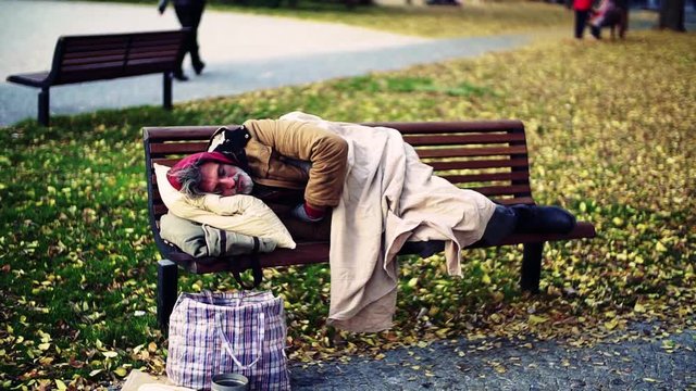 Homeless beggar man with a pillow lying on bench outdoors in city, sleeping.