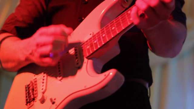A man strums his electric guitar to the rhythm of the beat at a live event, using his fingers and a plectrum to stump the guitar as the lights shine across the body of the guitar 