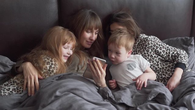 Mum with long brown hair lying together with her children in large grey bed. She showing them sth on the phone and children starring there. Small boy touching sister's nose. Family concept