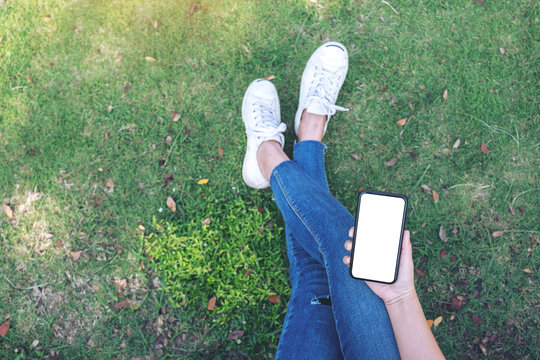 Top view mockup image of a woman holding black mobile phone with blank white screen while sitting in the outdoors