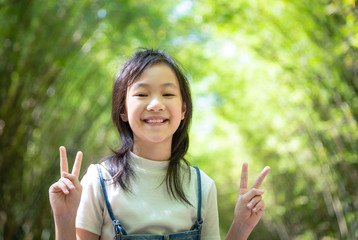 Portrait of smiling little asian girl shows a gesture of two fingers on bamboo forest,green nature...