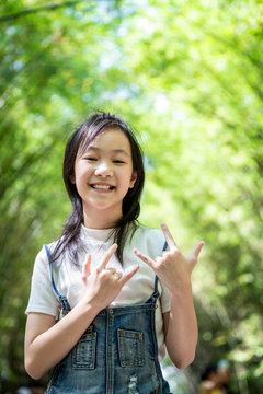 Portrait of smiling little asian girl right sign language meaning I Love You and look at the camera on bamboo forest,green nature background,happy people concept