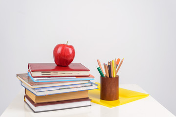 red juicy apple lies on a pile of training books on the table, beside it there are multicolored pencils, pens and markers in the stand and the yellow folder lies
