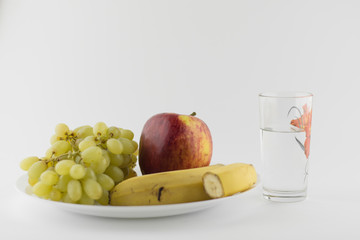 vegetarian still life: red ripe apples, bananas and seedless grapes on a white plate, close to the glass with pure water