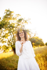 Caucasian happy girl in a white dress with a wreath of wild flowers in the summer at sunset in the field.