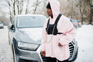 Stylish urban style african american man in pink hoodie posed against suv car at winter. Afro rapper guy.