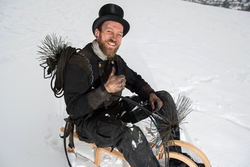 Daumen hoch, Schornsteinfeger in Arbeitskleidung und Kehrgerät sitzt auf Holzschlitten im Schnee