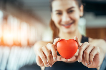 Asian sport woman holding red heart in fitness or gym center. Medical cadio heart strength training lifestyle. Pretty female sport girl workout exercise. Red massage ball in hand.