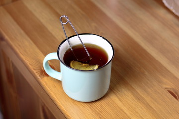 Hot warming tea with lemon in a light blue mug with a brewed tea strainer on a table with a wooden...
