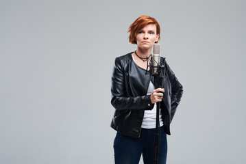 Portrait of rock singer wearing leather jacket and keeping static mic, sings a song loudly on grey...