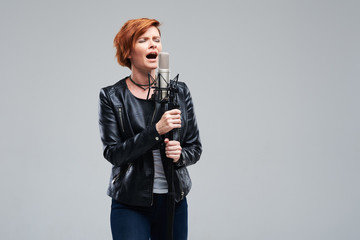 Portrait of rock singer wearing leather jacket and keeping static mic, sings a song loudly on grey...