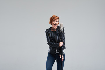 Portrait of rock singer wearing leather jacket and keeping static mic, sings a song loudly on grey...