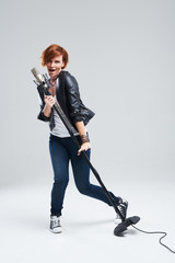 Fototapeta premium Full-length portrait of rock singer wearing leather jacket and keeping static mic, sings a song loudly on grey background. Concept of rock music and rave