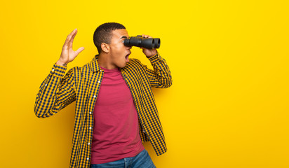 Young afro american man on yellow background and looking in the distance with binoculars