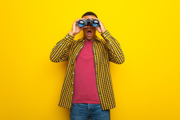 Young afro american man on yellow background and looking in the distance with binoculars