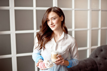 Beautiful young woman with dark hair dressed in a white shirt is standing in a cafe indoors with a glass cup of latte on soy milk.