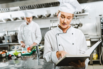 female chef in uniform reading recipe book while colleague cooking on background in restaurant kitchen