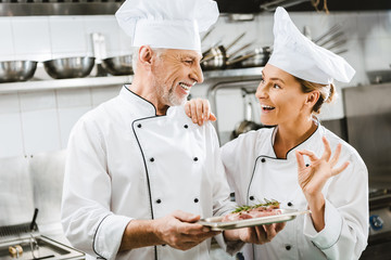 male chef presenting meat dish to smiling colleague showing ok sign in restaurant kitchen
